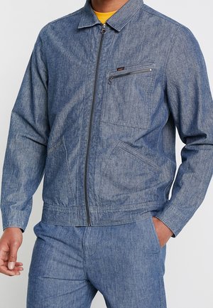 Veste en denim avec une finition texturée bleue, dotée d'une fermeture éclair à l'avant, de poches latérales et d'une poche poitrine avec une fermeture éclair.