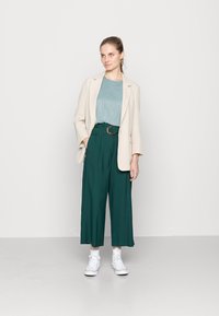 Blazer xadrez creme sobre uma blusa verde-água, calças largas verdes e sapatilhas brancas. Detalhes de bolsos e um cinto castanho nas calças. Texturas suaves.