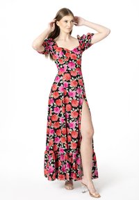 Robe maxi en tissu noir avec un motif floral vibrant en rose et rouge, manches bouffantes, décolleté carré et fente haute jusqu'à la cuisse. Sandales à talons transparents.