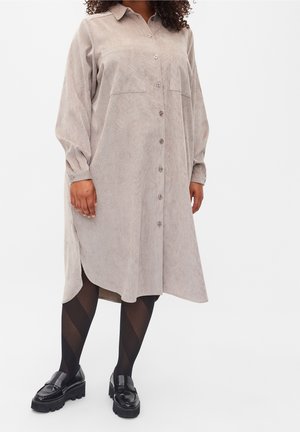 Kobieta ubrana w beżową, zapinaną na guziki, długorękawową sukienkę midi, wzorzyste czarne rajstopy i czarne masywne loafersy, stojąca na białym tle.