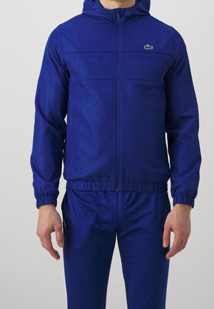 Hombre vistiendo una chaqueta con capucha y cremallera azul y pantalones a juego con logotipos de Lacoste en el pecho y el muslo.