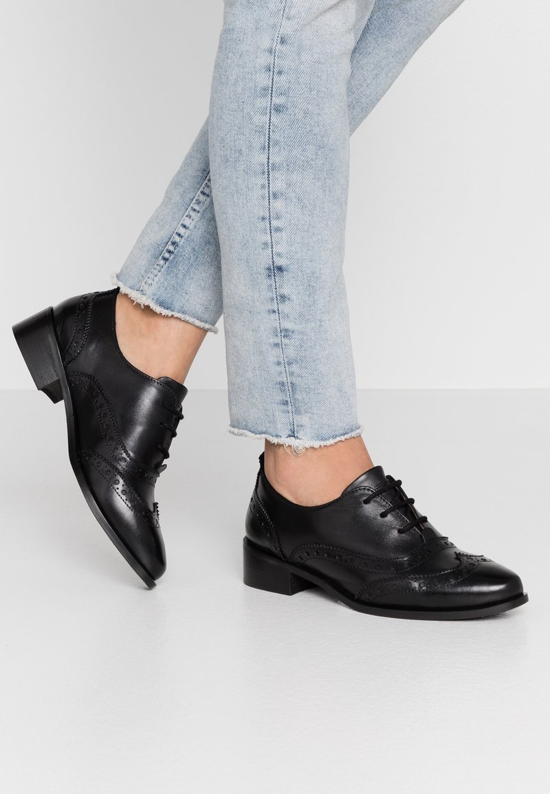 Chaussures Oxford en cuir noir avec un design texturé à bout aile, talon bloc bas et lacets à l'avant, associées à un jean court en denim clair.