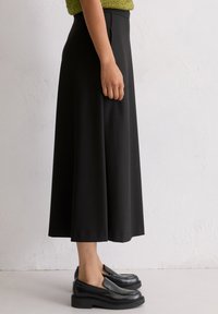 Schwarze, knielange Rock aus glattem Stoff mit einem taillierten Bund und sanfter A-Linie. Kombiniert mit schwarzen Slippern.