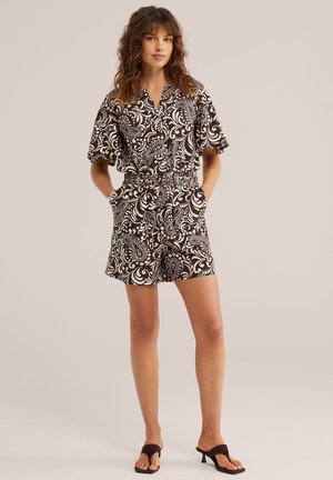 Žena s kučeravými vlasmi, oblečená v krátkom overale s čierno-bielym vzorom a čiernych sandáloch na opätku, stojaca s rukami v náprsných vreckách.