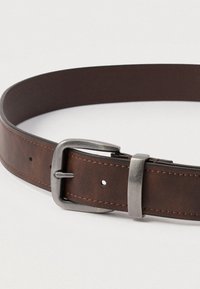 Καφέ δερμάτινη ζώνη με ασημένια μεταλλική αγκράφα και θηλιά, δοσμένη επίπεδη σε λευκή επιφάνεια με ορατές ραφές και τρύπες ρύθμισης.