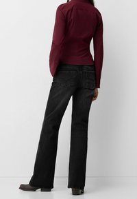 Camicia fitted bordeaux a maniche lunghe, abbinata a jeans neri a zampa d'elefante. Presenta chiusure con bottoni e dettagli sottili sulle tasche posteriori.