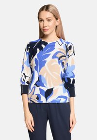 Pull en motif floral bleu avec des manches trois-quarts, fabriqué à partir d'un matériau doux, présentant différentes nuances de bleu, des accents crème et noir.