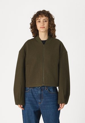 Cropped olive green zip-up jacket with a high collar, loose long sleeves, and soft fabric texture, paired with blue denim jeans.