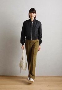 Giacca bomber nera con zip, pantaloni larghi verde oliva, borsa bianca e sneakers. Modella cammina su un pavimento di legno contro un muro grigio.
