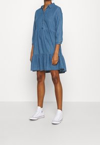 Jeansklänning med knäppt placket, krage, trekvartsärmar och volangkjol i flera lager. Buren med vita högstövlar.