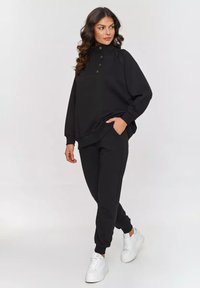 Czarna bluza z wysokim kołnierzem, zapięciem na guziki i luźnym krojem, w zestawie z dopasowanymi spodniami i białymi sneakersami. Miękka tkanina, minimalistyczny design.