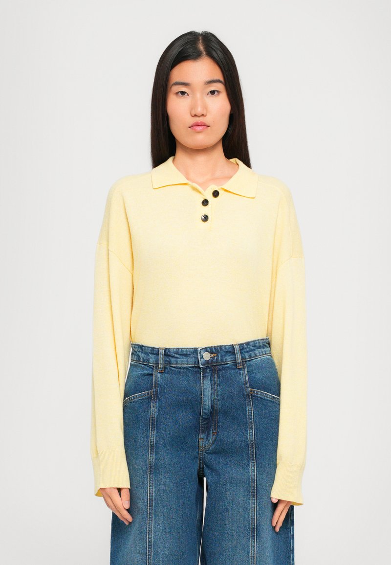 Woman with long black hair wearing a pale yellow long-sleeve polo shirt and high-waisted wide-leg blue jeans standing against a plain background.