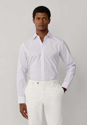 Mann trägt ein weißes und hellrosa kariertes Hemd mit langen Ärmeln, das in weiße Hose gesteckt ist, mit einer Hand in der Tasche, steht vor einem einfarbigen Hintergrund.