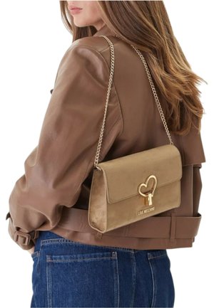 Béžová kabelka se zlatým srdcovým zapínáním a řetízkovým popruhem, s semišovou strukturou. N nošená přes rameno s hnědou bundou.