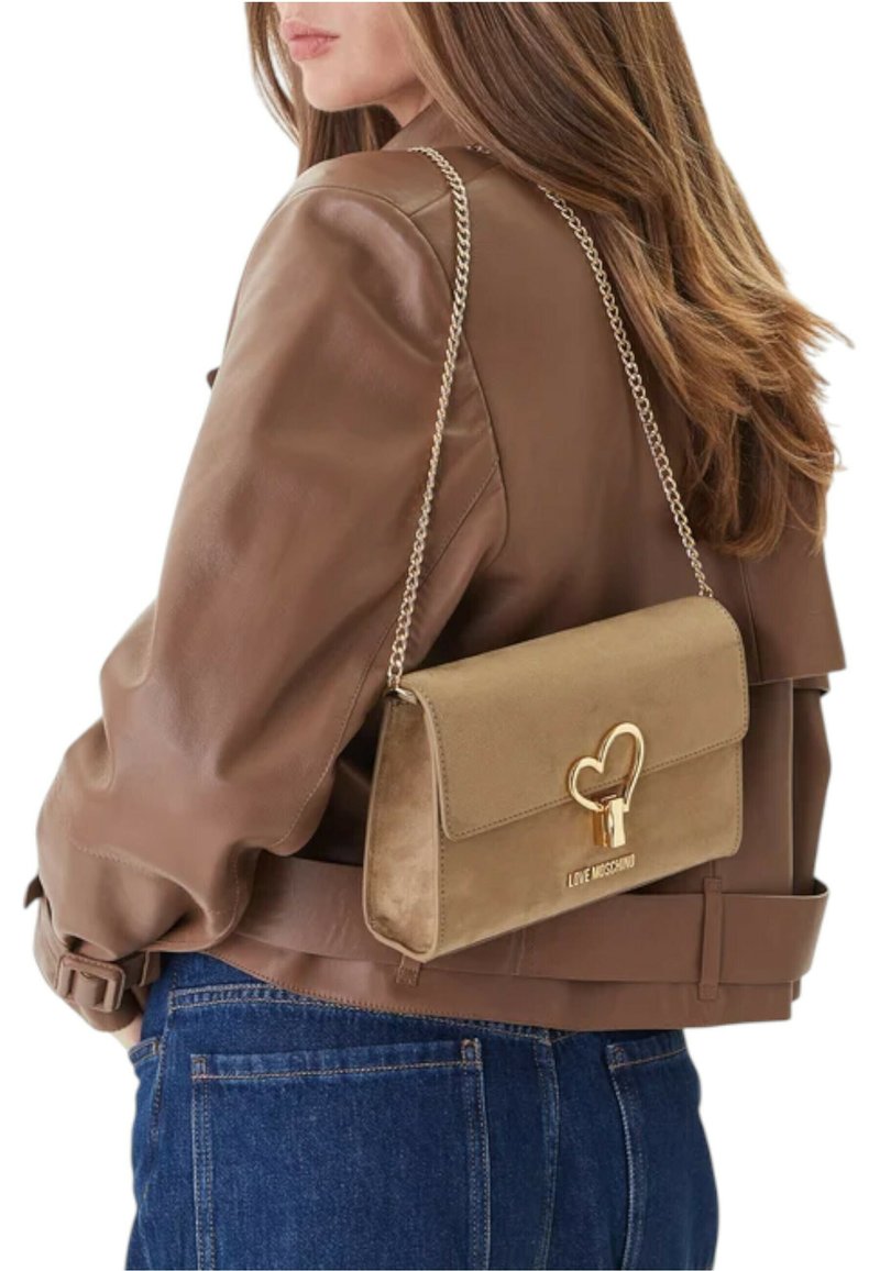 Beige käsilaukku, jossa on kultainen sydämenmuotoinen lukko ja ketjuhihna, mokkamainen pinta. Käytetty olkapäällä ruskean takin kanssa.