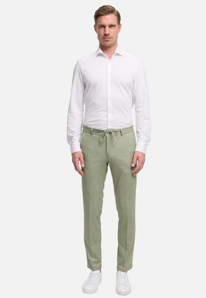 Man standing wearing a white button-up shirt, light green drawstring trousers, and white sneakers on a plain light background.