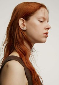 Pendientes plateados redondos con piedras transparentes, llevados en la oreja de una persona con cabello largo y ondulado de color rojo, sobre un fondo neutro.