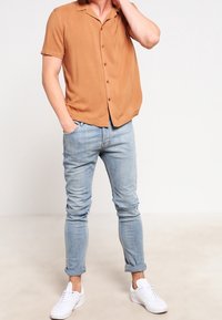 Kortärmad, kragskjorta i brunt tyg, parat med ljusblå, figursydda jeans och vita sneakers. Enkla knappdetaljer, avslappnad passform.