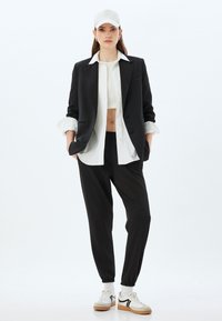 Zwart tailored blazer, witte overhemdbloes, cropped top, zwarte joggers en witte sneakers met zwarte accenten; lichtgrijze pet.