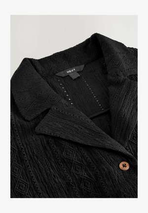 Schwarze strukturierte Jacke mit steigendem Kragen, ausgestattet mit einem Rautenmuster, einem einzelnen braunen Knopf und sichtbaren Nähten.