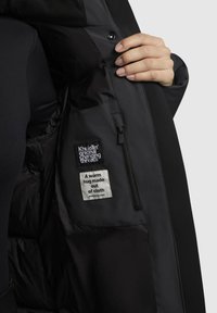Interno della giacca nera caratterizzato da un materiale liscio, una tasca con zip e due etichette in tessuto con la scritta "original changing threads" e "Un abbraccio caldo fatto di stoffa."