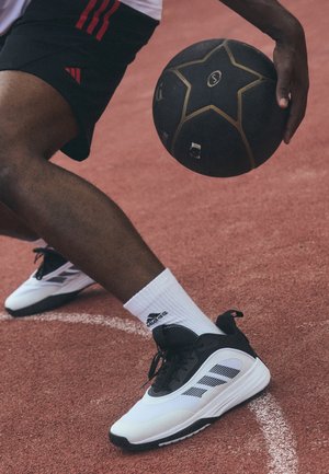 Person, der dribler med en sort og gul basketball på en rød bane, iført hvide Adidas-sneakers og sorte shorts med røde striber.