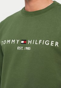 Green sweatshirt made of soft material featuring white embroidered text "TOMMY HILFIGER" and "EST. 1985" with a rectangular logo in red, white, and blue.