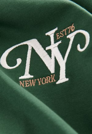 Vihreä kangas, jossa on valkoisella kirjailtuna kirjaimet "NY" ja "NEW YORK", sekä "EST 76" vaaleanruskealla, esitellen teksturoitua muotoilua.