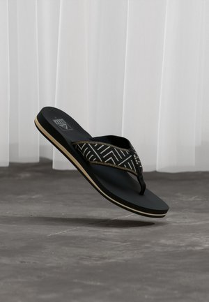 Zwarte teenslipper met beige zoolrand en bandje van stof met geometrisch patroon, zwevend boven een betonnen vloer en een witte gordijnachtergrond.