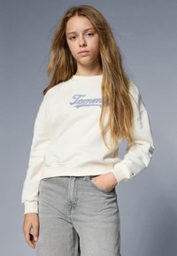 Hanorac alb cu manșete cu nervuri, având logo-ul brodat "Tommy" multicolor. Asortat cu blugi gri, expus pe un fundal gri.