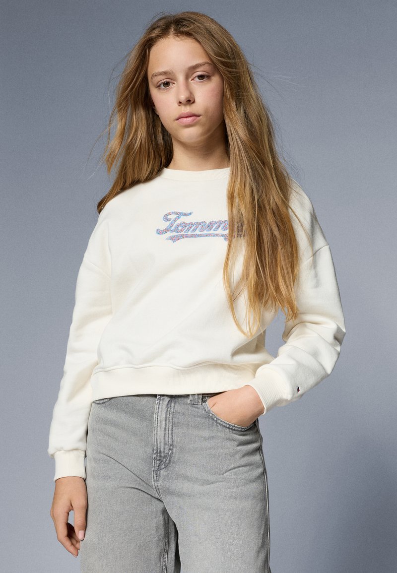 Hanorac alb cu manșete cu nervuri, având logo-ul brodat "Tommy" multicolor. Asortat cu blugi gri, expus pe un fundal gri.