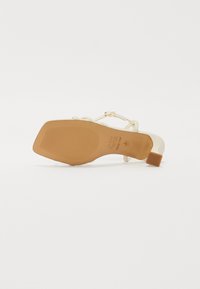 Sandale à talons avec une tige crème clair, des sangles texturées, une boucle réglable et une semelle en caoutchouc marron présentant des lignes horizontales.