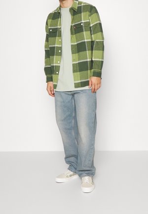 Grünes kariertes Flanellhemd mit zwei Brusttaschen, getragen über einem hellgrünen T-Shirt; hellblaue Jeans mit lockerem Schnitt und gemusterte weiße Sneakers.