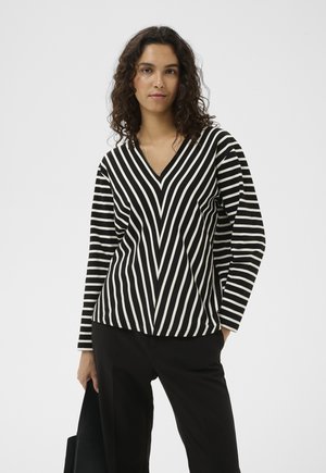 Žena s kučeravými vlasmi, oblečená v čierno-bielom pruhovanom tričku s výstrihom do V a čiernych nohaviciach, držiaca v jednej ruke čierny kabát.