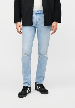 Mężczyzna ubrany w jasnoniebieskie podarte dżinsy, czarną kurtkę, białą koszulę oraz czarne trampki z białymi paskami, stojący na jednolitym tle.