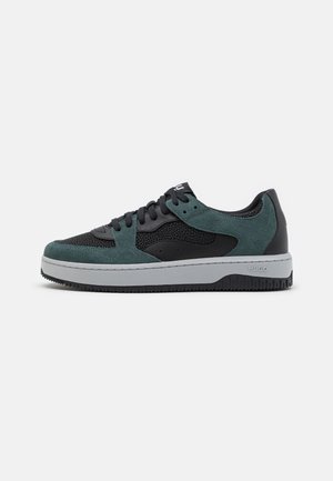 Low-top sneaker met zwarte mesh en groene suède overlays, zwarte veters en een dikke witte zool met "Hugo" branding op de hiel.