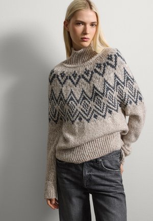 Beiger Strickpullover mit einem dunkelblauen geometrischen Muster, mit hohem Kragen und gerippten Bündchen, kombiniert mit dunklen Jeans.