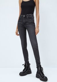 Person med svart ärmlös topp, mörkgrå skinny jeans och svarta chunky snörning ankla stövlar som står mot en enkel ljus bakgrund.