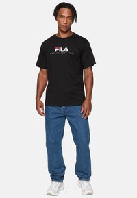 Sort bomulds T-shirt med hvid og rød "FILA" logo og teksten "ETABLERT 1911", parret med blå denim jeans og hvide sneakers.