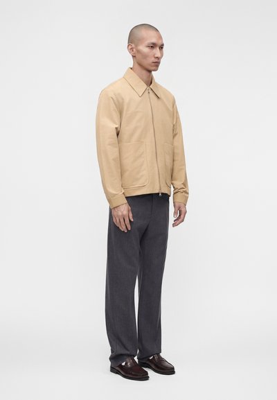 Homme debout portant une veste beige zippée avec de grandes poches avant, un pantalon gris foncé et des chaussures en cuir marron foncé.