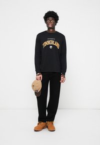 Crna majica dugih rukava s "Authentic Timberland" logotipom u žutoj boji, crne hlače, smeđe čizme i smeđi šešir u ruci.