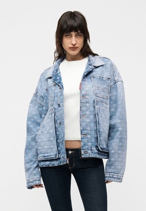 Jonge vrouw die een lichtblauwe oversized spijkerjack met patroonachtige textuur draagt over een witte cropped top en donkere spijkerbroek, staand tegen een effen achtergrond.