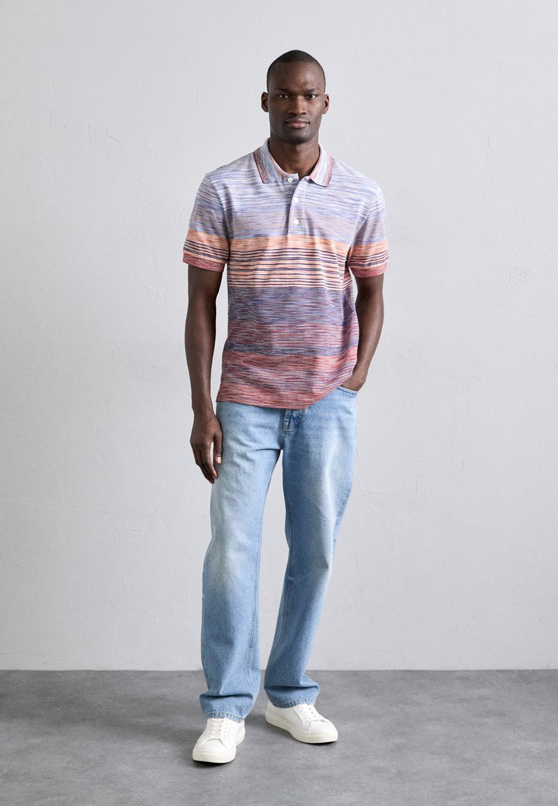 Gestreiftes Polohemd mit blauen, orangefarbenen und lila horizontalen Linien. Getragen mit hellblauen Jeans und weißen Turnschuhen. Kurzärmlig, mit Kragen.