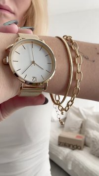 Arany óra fehér tárcsával, hálós szíjjal, minimalista órás jelzőkkel, kísérőként két arany karkötővel. A háttér semleges dekorációval rendelkezik.