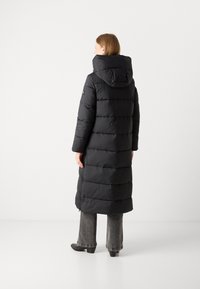 Svart lang pufferjakke med hette, kviltet design, horisontale seksjoner og sidelommer med glidelås. Båret over grå flarebukser og svarte sko.