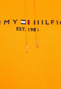 Sudadera amarilla con cordones con puntas plateadas. Texto en negrita que dice "TOMMY HILFIGER EST. 1985," con un logo en rojo, blanco y azul.