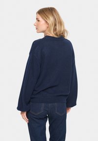 Mornarska pletena jopica z udobnim krojem, okroglim izrezom in zapletenimi detajli na ramenih. Združena z temnimi jeans hlačami. Mehka tekstura.