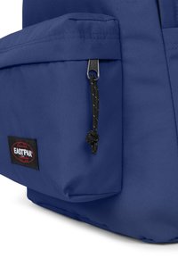Zaino in tessuto blu con tasca frontale dotata di zip e tirazip nero, che mostra il patch del logo Eastpak rosso e nero.