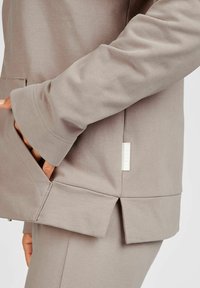 Jasnobrązowa bluza z długimi rękawami, bocznymi kieszeniami oraz rozcięciem na dole; posiada małą metkę marki na dolnym boku.