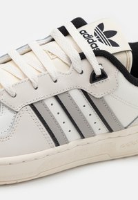 White leather sneaker with three grey stripes, black accents, and white laces. Features a textured sole and an adidas logo on the tongue.